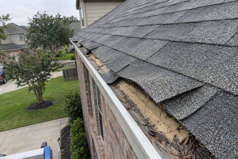 Shingle tabs lifted along a roof rake edge with bare adhesive strip and dirt underneath