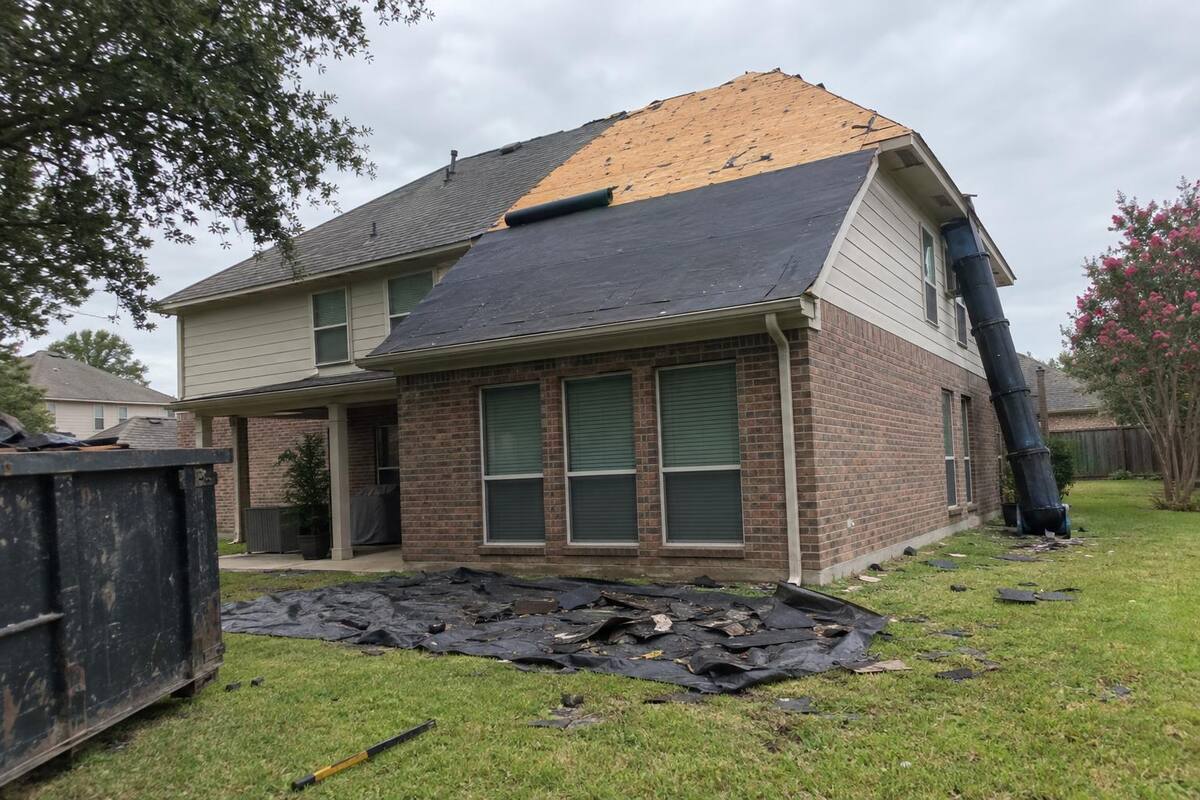 Tear-off in progress on a two-story brick-veneer house with old shingles stripped from one slope and underlayment exposed
